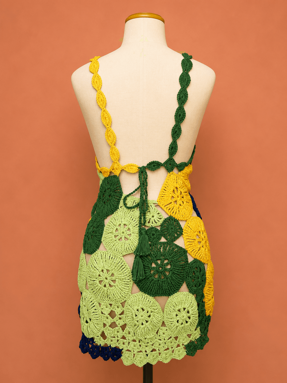 Vestido Tarsila de crochê artesanal patchwork colorido, confeccionado à mão com motivos circulares em tons de azul, verde, amarelo e verde-claro. A peça possui modelagem curta, alças trabalhadas em crochê e design vazado, apresentada em manequim off white sobre fundo terracota suave, destacando a riqueza dos pontos e o acabamento artesanal. Ideal para festas, eventos, produções de verão e ocasiões especiais.