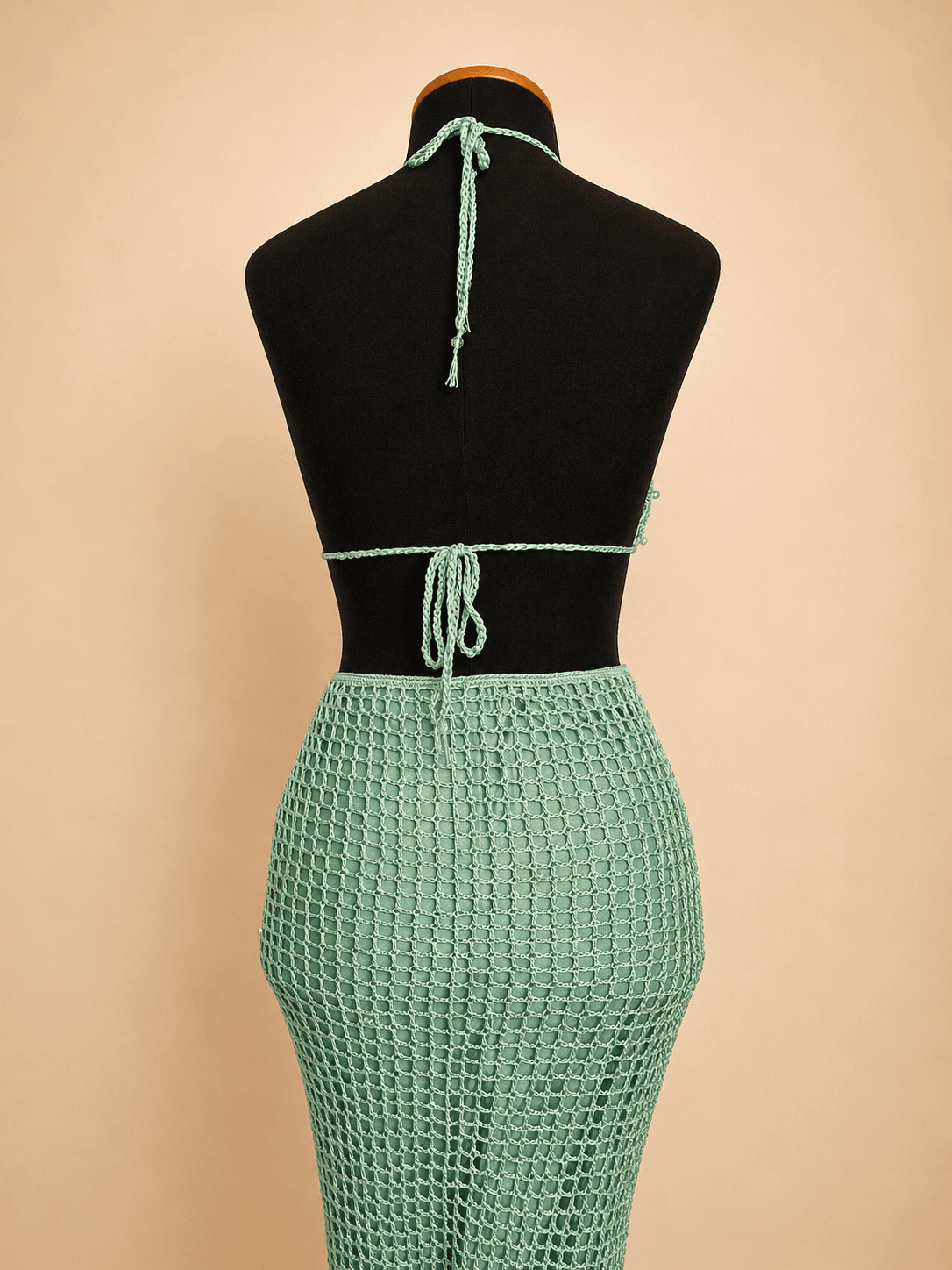 Vestido longo de crochê artesanal na cor verde mint, composto por top com decote triangular, recorte na cintura com argola metálica central e saia longa em trama vazada. A peça possui delicadas aplicações de miçangas e está exposta em um manequim preto, destacando os detalhes do acabamento e da modelagem sereia. Ideal para festas, shows, eventos de verão e produções à beira-mar.