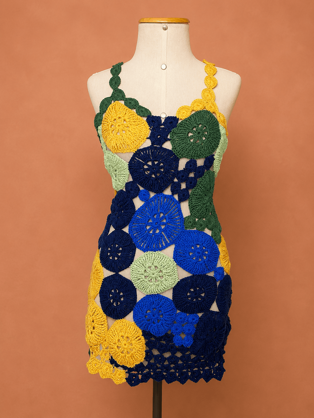 Vestido Tarsila de crochê artesanal patchwork colorido, confeccionado à mão com motivos circulares em tons de azul, verde, amarelo e verde-claro. A peça possui modelagem curta, alças trabalhadas em crochê e design vazado, apresentada em manequim off white sobre fundo terracota suave, destacando a riqueza dos pontos e o acabamento artesanal. Ideal para festas, eventos, produções de verão e ocasiões especiais.