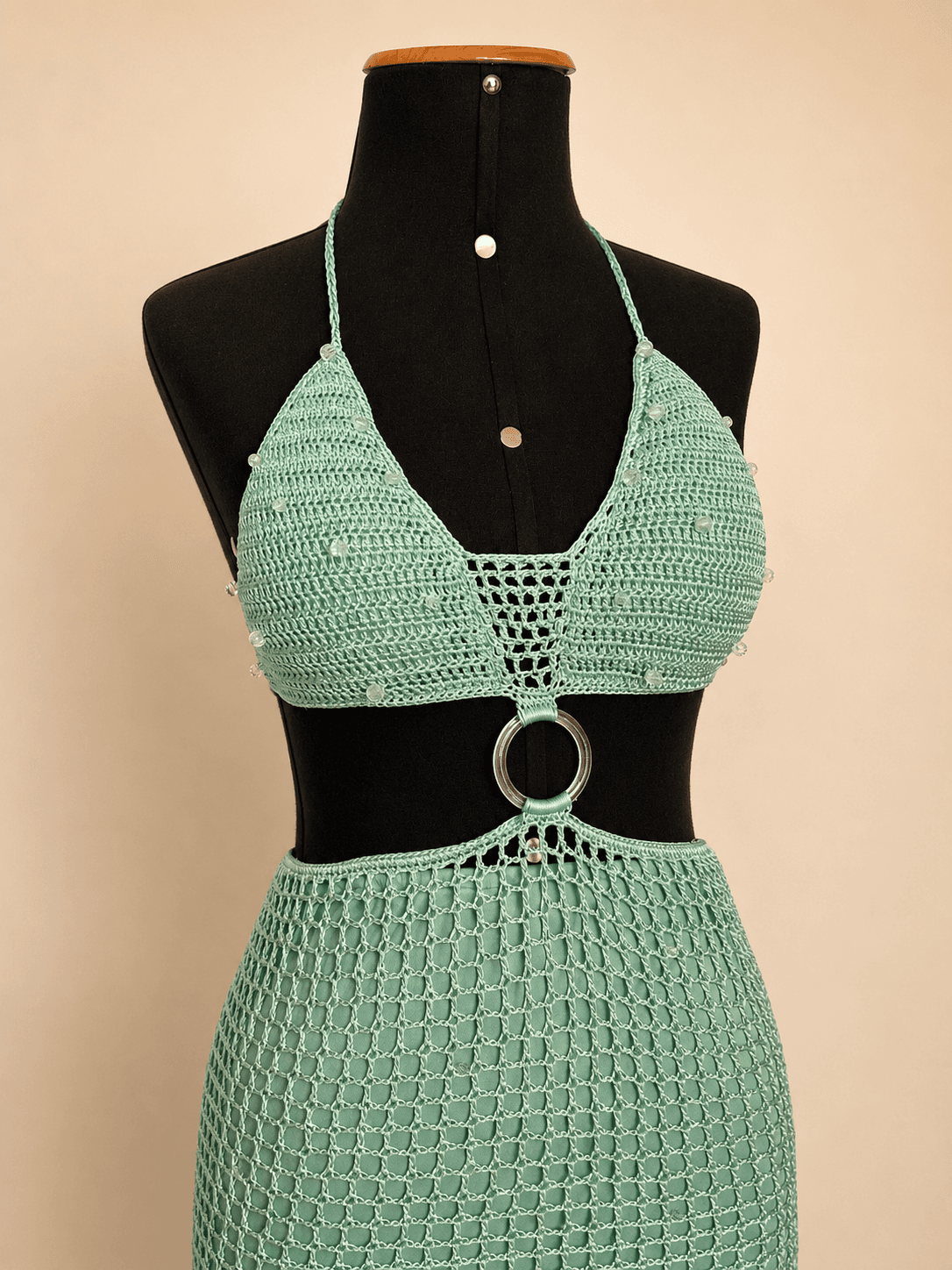 Vestido longo de crochê artesanal na cor verde mint, composto por top com decote triangular, recorte na cintura com argola metálica central e saia longa em trama vazada. A peça possui delicadas aplicações de miçangas e está exposta em um manequim preto, destacando os detalhes do acabamento e da modelagem sereia. Ideal para festas, shows, eventos de verão e produções à beira-mar.