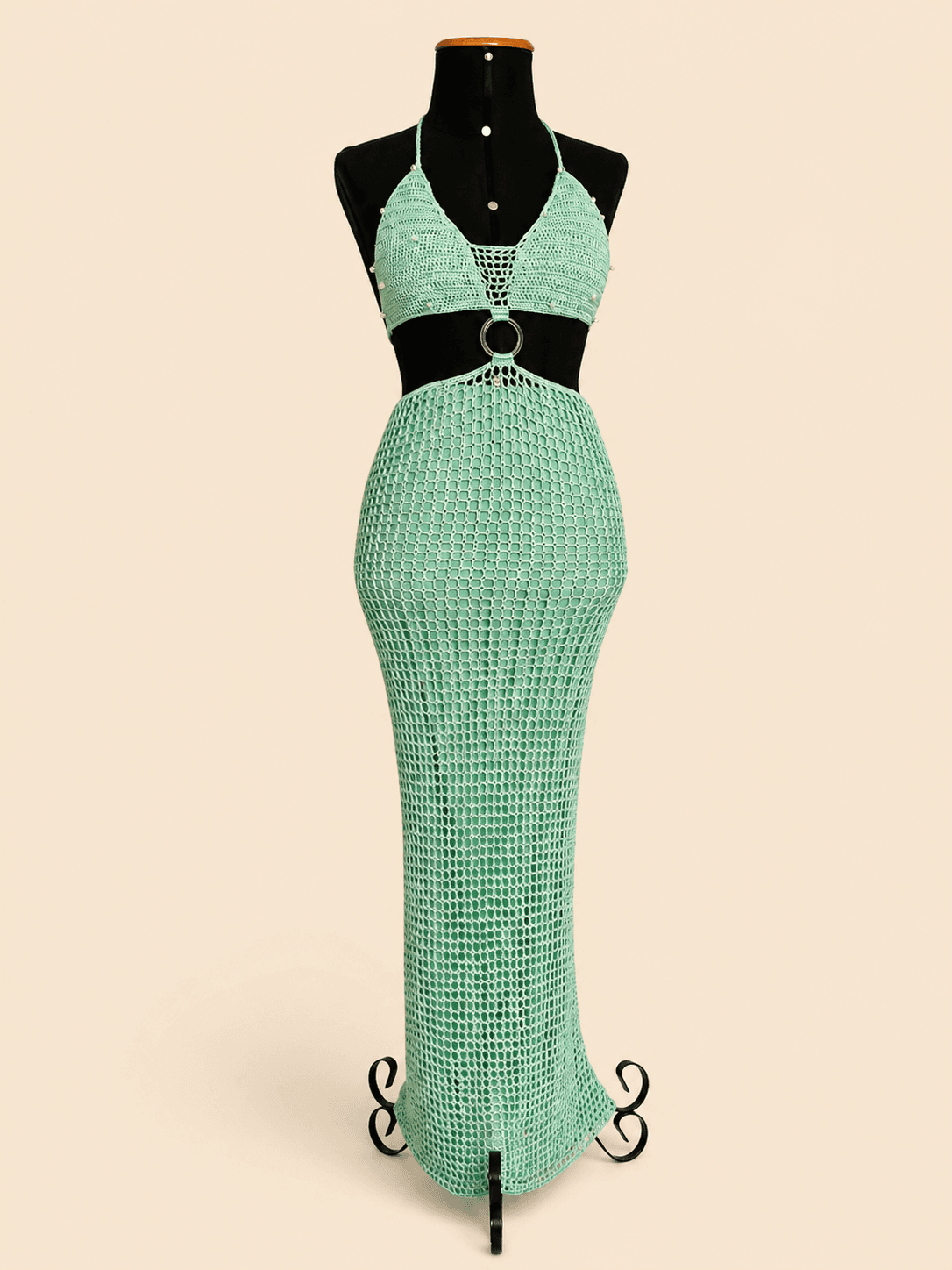 Vestido longo de crochê artesanal na cor verde mint, composto por top com decote triangular, recorte na cintura com argola metálica central e saia longa em trama vazada. A peça possui delicadas aplicações de miçangas e está exposta em um manequim preto, destacando os detalhes do acabamento e da modelagem sereia. Ideal para festas, shows, eventos de verão e produções à beira-mar.